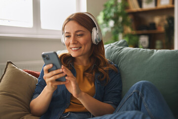 A woman with long hair smiles while sitting comfortably on a couch with headphones on. She is engaged with her smartphone in a bright and inviting living room filled with plants.