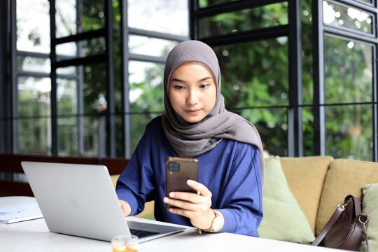 Young Asian Muslim Business Woman Wearing Hijab using Smartphone with Laptop at a Cafe