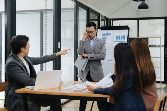 Conflict and Decisions in Boardroom: The photo captures a tense moment in a modern office, with a man pointing his finger at a colleague during a business meeting, and others engrossed in documents.