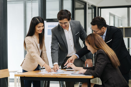 Collaborative Business Meeting: Four diverse professionals gather around a table, reviewing paperwork and exchanging ideas in a modern office setting, highlighting teamwork and strategic planning.