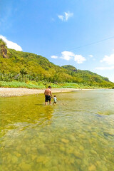 Happy Asian father and daughter enjoy playful in river with hills in background. Happy family time, outdoor adventure, bonding moment, rural nature, childhood joy, and summer activity
