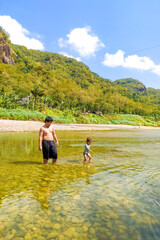 Happy Asian father and daughter enjoy playful in river with hills in background. Happy family time, outdoor adventure, bonding moment, rural nature, childhood joy, and summer activity