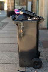 Overflowing trash can on a quiet city street showcases the need for better waste management and environmental awareness at the start of a sunny day