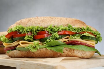 Tasty baguette sandwich with smoked sausage, cheese and vegetables on wooden board against grey...