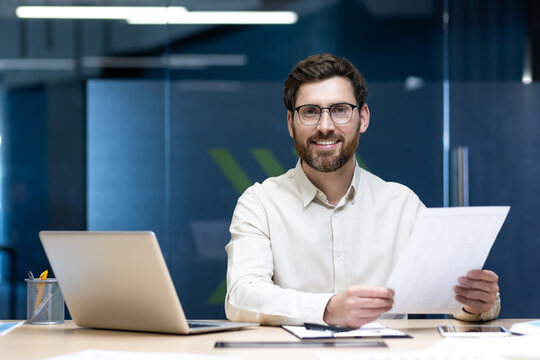 Portrait of a smiling and successful young businessman sitting at a desk in an office center, working on a laptop and with documents