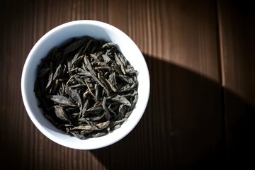 White bowl filled with dry tea leaves on a dark wooden surface lit by light creating a strong shadow