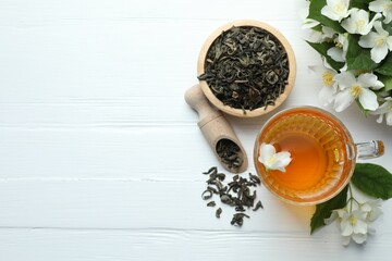 Aromatic jasmine tea in glass cup, flowers and leaves on white wooden table, flat lay. Space for...