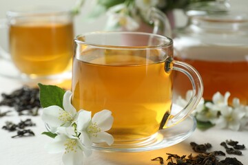 Aromatic jasmine tea in glass cup, teapot, flowers and dried leaves on white table, closeup