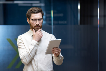 A serious young man is standing in the office and staring intently at the screen of the tablet he...