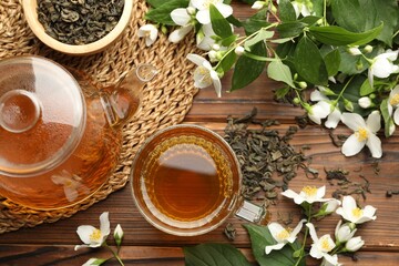 Aromatic jasmine tea in glass cup, teapot, brew and flowers on wooden table, flat lay