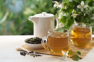 Tasty jasmine tea in cups, teapot, dry leaves and flowers at white wooden table, closeup. Space for text