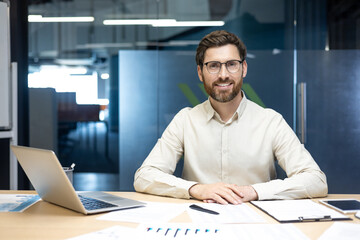 Portrait of a young smiling man in a shirt and glasses sitting in the office at a table with a...