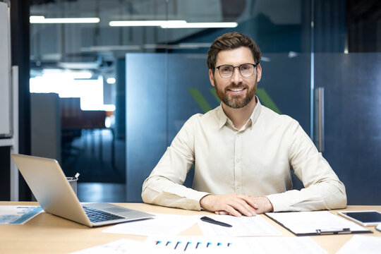 Portrait of a young smiling man in a shirt and glasses sitting in the office at a table with a laptop and documents, looking at the camera
