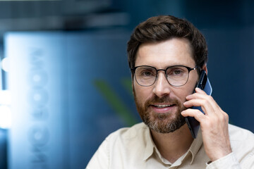 Close-up portrait of a young male businessman in the office, talking on the phone and looking at...