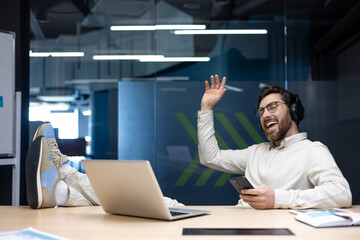 Relaxed young man sitting at office desk wearing headphones, listening to music and gesturing with...