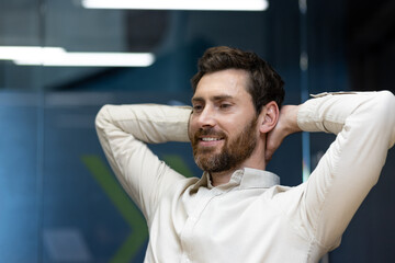 Close-up photo of a young smiling man sitting in the office, hands behind his head, resting and...
