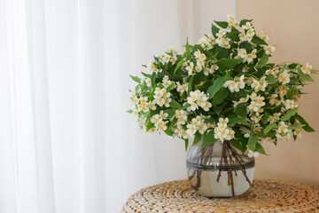 Beautiful jasmine flowers in vase on wicker table near window indoors, space for text
