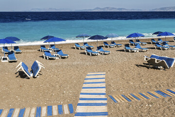 sand beach with blue deck chairs and umbrellas arranged in rows. A wooden walkway leads from the...