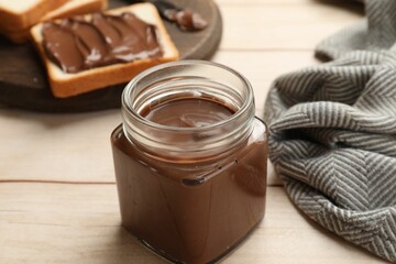 Tasty chocolate butter in glass jar and slices of bread on light wooden table, closeup