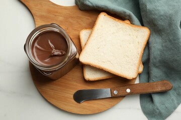 Tasty chocolate butter in glass jar, slices of bread and knife on white marble table, top view