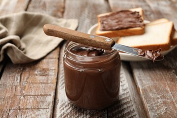 Tasty chocolate butter in glass jar and knife on color wooden table, closeup