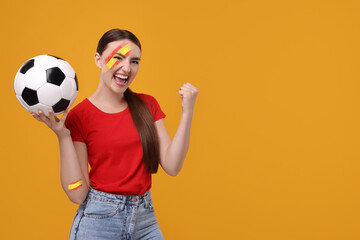 Excited fan with colors matching Spain flag on her face and arm holding soccer ball against orange...