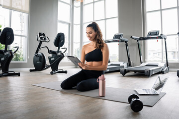 A woman is sitting on a mat in a gym with a tablet in her lap. She has a pink bottle next to her