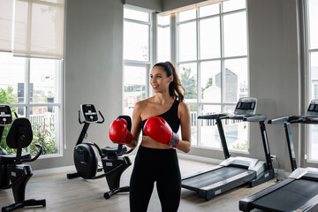A woman wearing a red boxing glove stands in front of a treadmill. She is smiling and she is...
