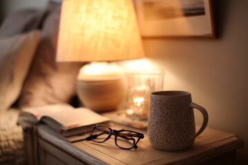 A cozy bedroom nightstand with a warm-toned lamp, a half-read book, a pair of reading glasses, and a ceramic mug, softly lit by ambient evening light.