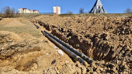 The pipes are laid in a trench dug towards the road with fencing and buildings. Nearby are piles of...