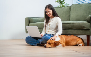 Asian young woman sits on the floor beside her sofa, gently stroking her Corgi dog head relax at...