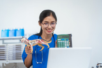 An Asian female veterinarian in blue scrubs and glasses works diligently at her desk, using a canine anatomical model and laptop to diagnose and plan treatment in a professional clinical environment.