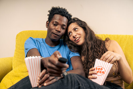 A happy diverse couple talking and choosing a movie to watch together on the sofa at home.