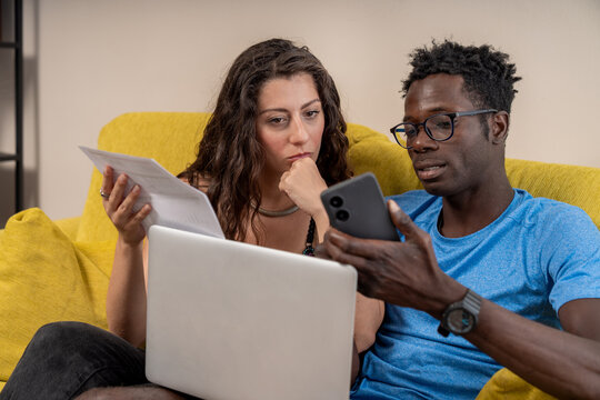A young interracial couple works from home or studies online, using a laptop together on their sofa.