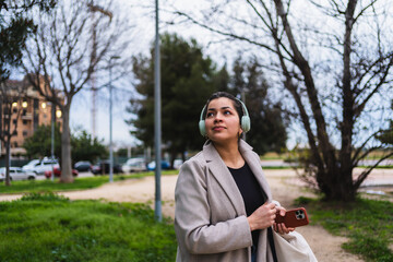 Young stylish businesswoman listening to music with headphones while walking in a park, holding her smartphone