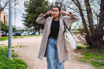Young stylish influencer adjusting headphones while listening to music on her smartphone and walking in a park