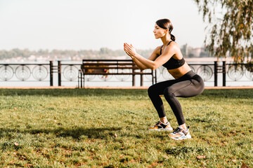Side view of athlete girl in stylish sportswear squatting exercising during workout on green park...
