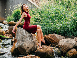 Young girl in luxurious red dress is sitting on a large rock by a mountain fast river, green bushes...