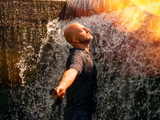 A bald man is standing under a waterfall, getting wet. Concept of relaxation and rejuvenation, as...