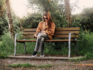 Young and stylish teenager girl sitting on a bench in a park alone. Waiting for date concept. The model wears trendy brown jacket and dark glasses and looks calm and elegant. Selective focus.