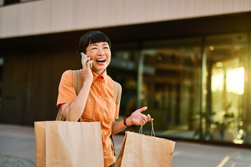 Portrait of a happy asian woman holding shopping bags, using a smartphone mobile phone and walking...