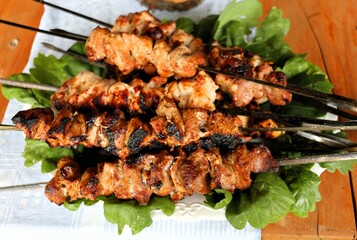 Juicy grilled pork skewers arranged on fresh green leaves atop a white plate. Traditional barbecue meal presented on a rustic wooden table for outdoor celebration.