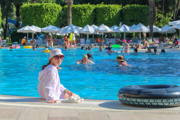 Kid girl in panama sits on edge of resort hotel pool with legs dangling in water