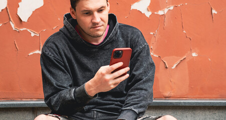 On a sunny day a young man wearing a gray hoodie and denim shorts is sitting on the pavement...