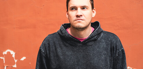 A young man wearing a gray hoodie stands with a serious expression against an orange wall in an...