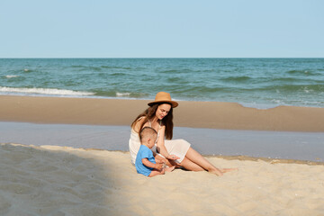 A loving mother in a white dress and hat sits on a sunny sandy beach, interacting with her baby in...