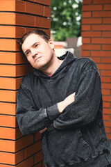 A man in a dark hoodie leans against a brick column lost in thought while standing in an outdoor...