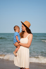 
A loving mother in a white dress and hat holds her baby boy on a sunny beach, gazing at him with...