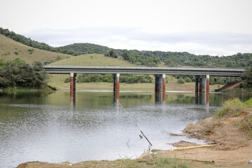 Foto de uma ponte cruzando uma represa em meio à natureza. As águas calmas refletem a estrutura e a vegetação ao redor, compondo uma cena tranquila e harmônica. Ideal para temas de infraestrutura, mei