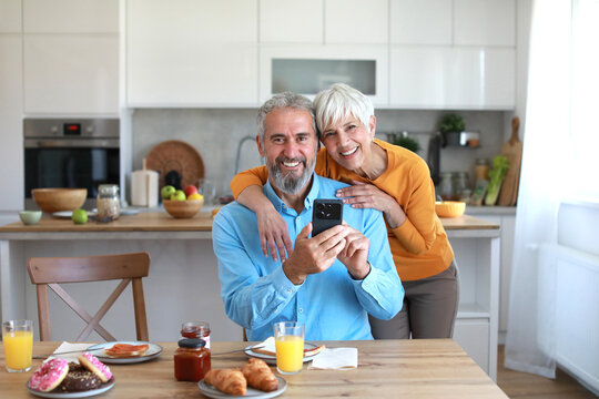 Portrait of an elderly senior couple having breakfast and using a smart phone at home. Happy healthy affectionate senior couple talking and showing smartphone screen to one another and texting and sit